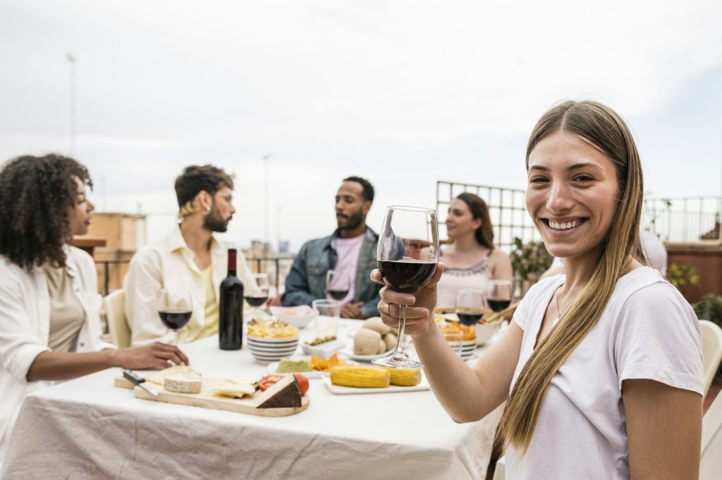 Young cheerful lady looking at camera with a glass of wine in a rooftop dinner party. Joyful